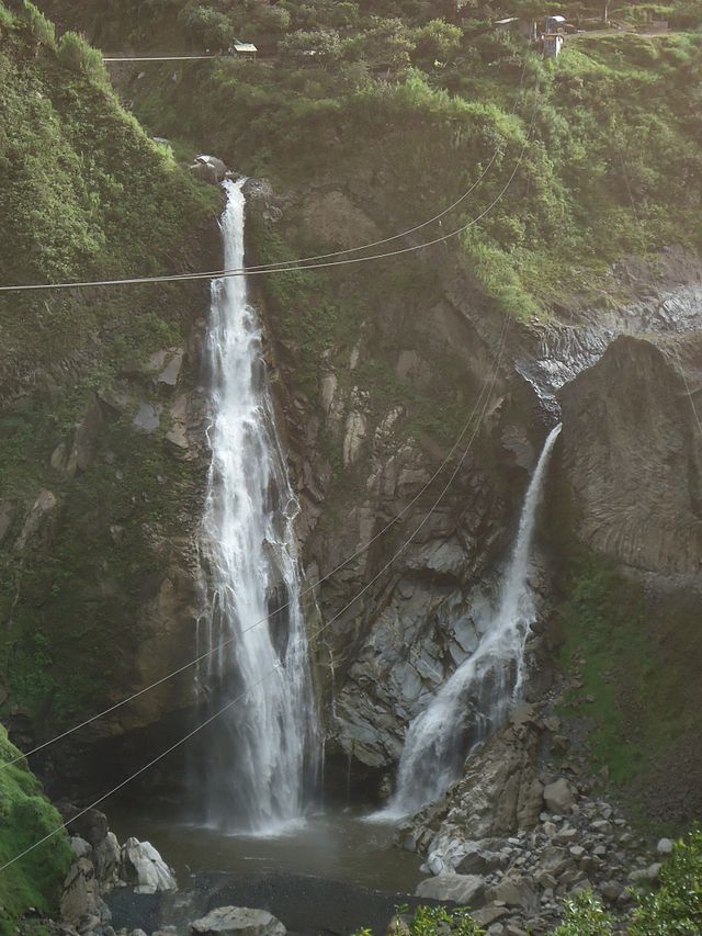 Zwei Wasserfälle stürzen eine felsige grüne Klippe hinab, Stromleitungen kreuzen