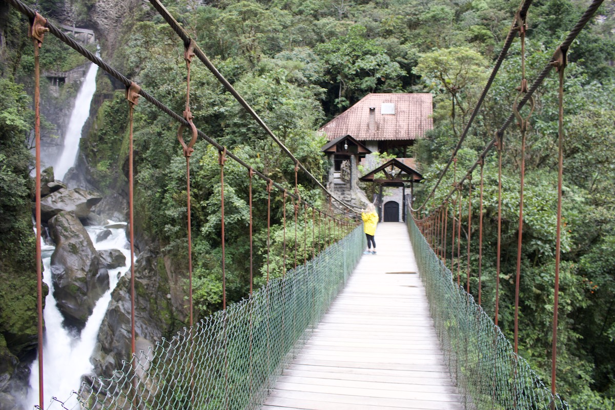 Suspension bridge over a forested gorge with a waterfall and a stone building