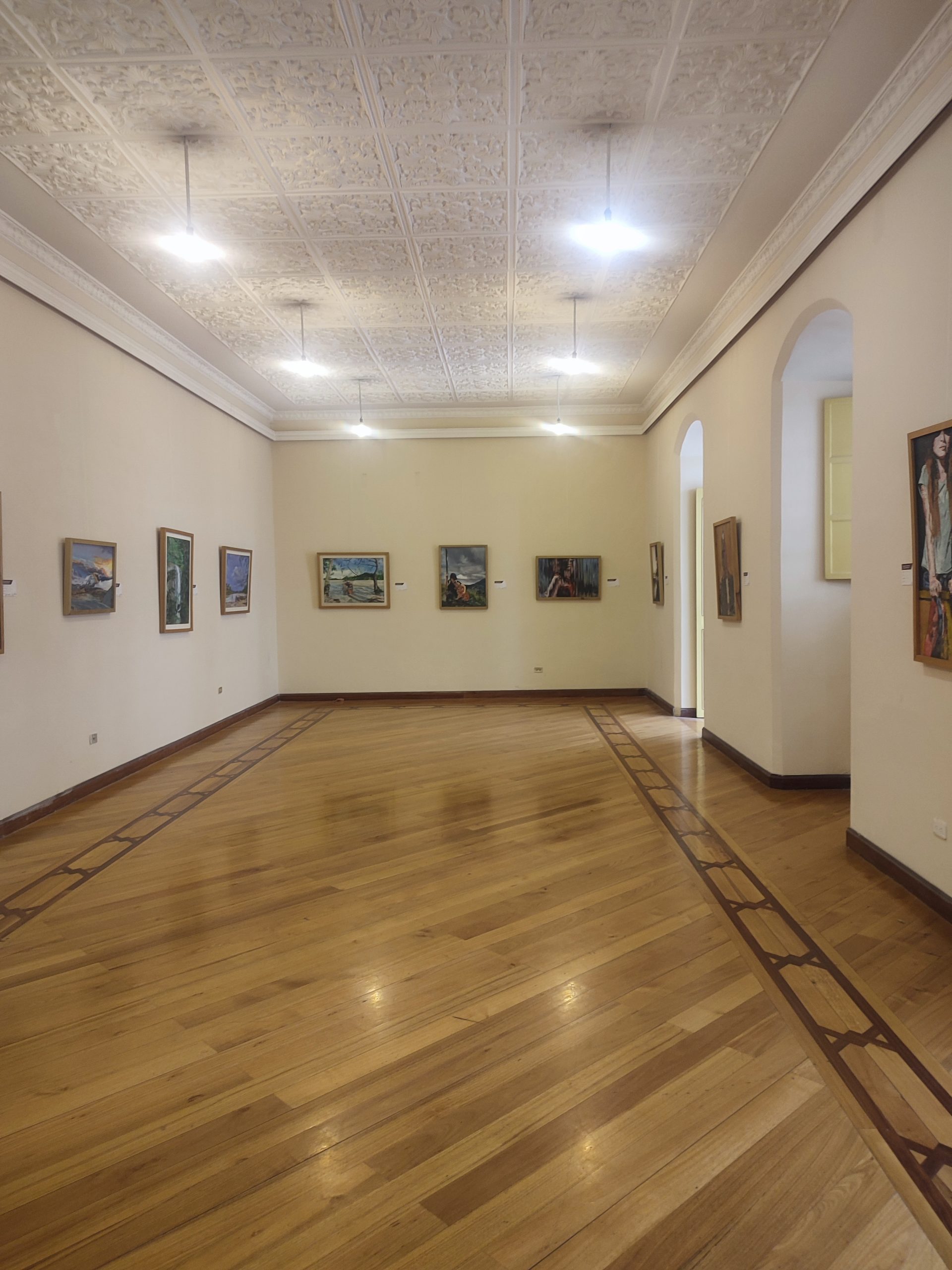 Galerieraum mit gerahmten Gemälden, Holzboden und verzierter weißer Decke