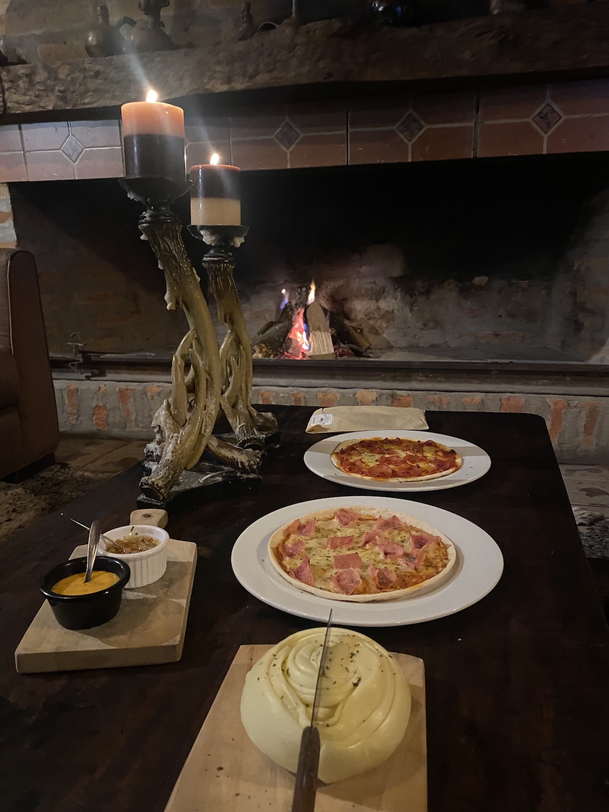 Dos pizzas pequeñas y un queso redondo sobre tablas frente a una chimenea encendida